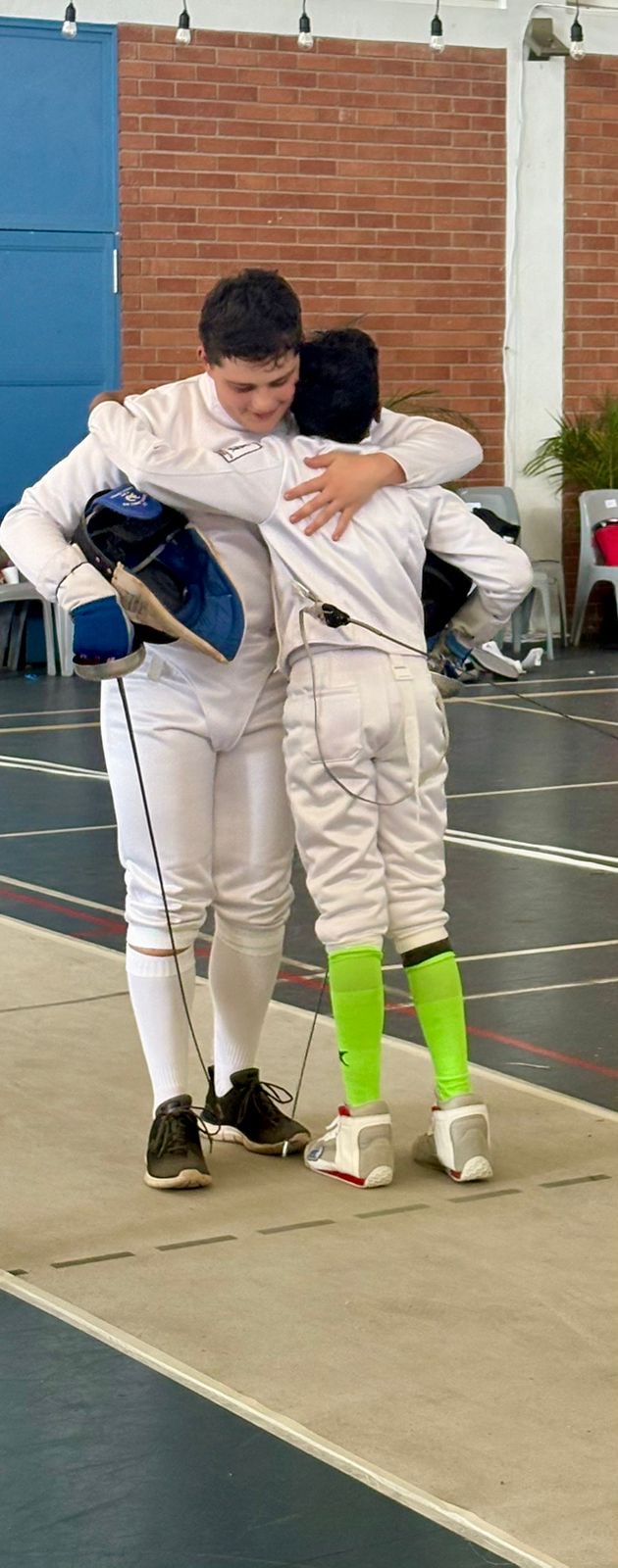 Two young fencers in full kit embracing on the piste after their bout