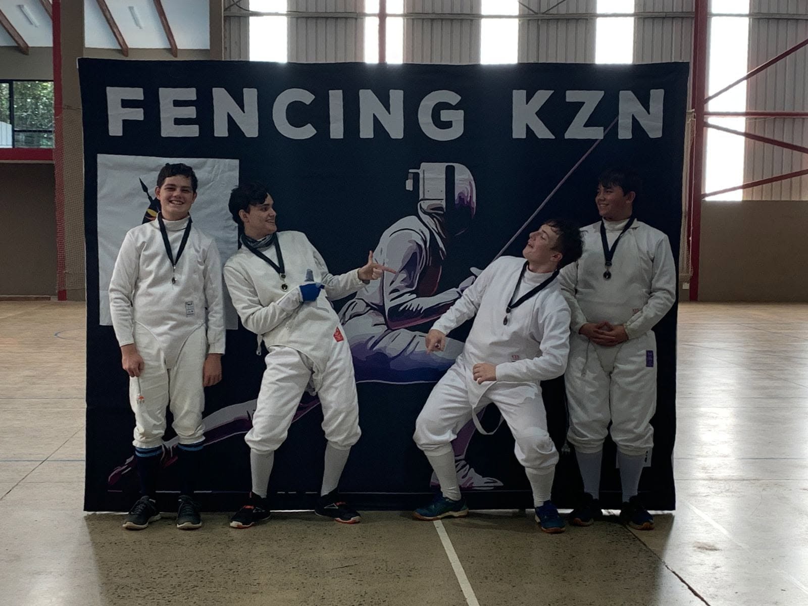 Four young fencers with medals joking around in front of the Fencing KZN banner